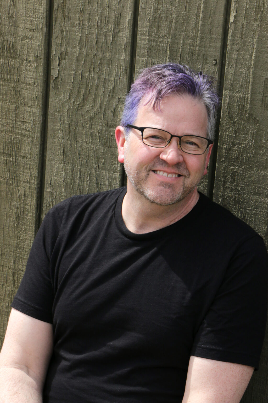 Smiling man with purple-tinted hair, glasses, and a black shirt, standing in front of a weathered wooden fence.