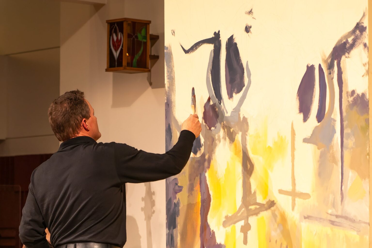 Man in a dark jacket painting a large abstract mural on a wall inside a gallery.