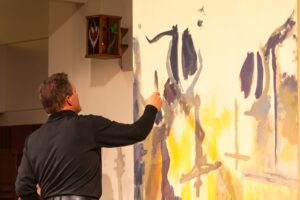 Man in a dark jacket painting a large abstract mural on a wall inside a gallery.