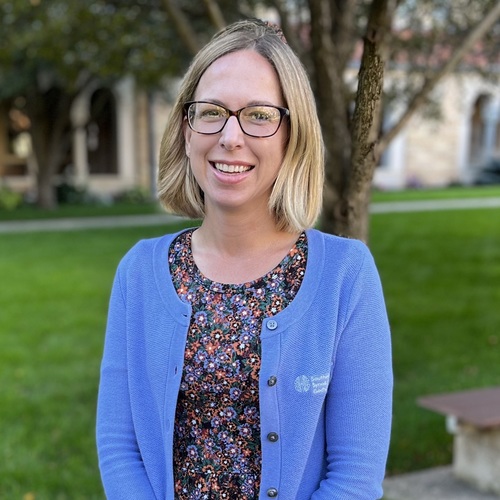 Smiling woman with blonde hair and glasses, wearing a blue cardigan, outdoors on a campus lawn.