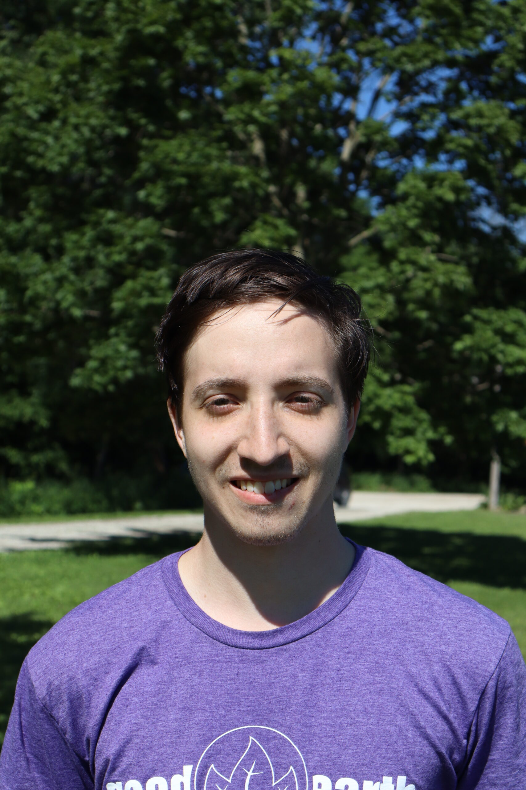 Young man with short dark hair smiling outdoors in a sunny park, wearing a purple shirt.