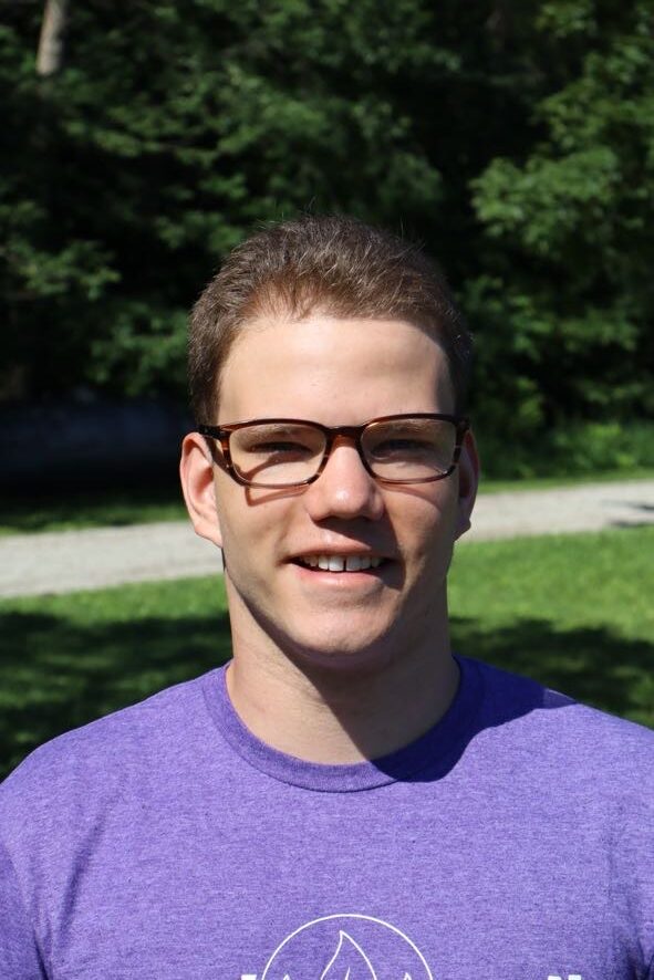 Young man with short brown hair and brown rectangular glasses smiles at the camera, wearing a purple T-shirt outdoors in a sunny park with green trees in the background.
