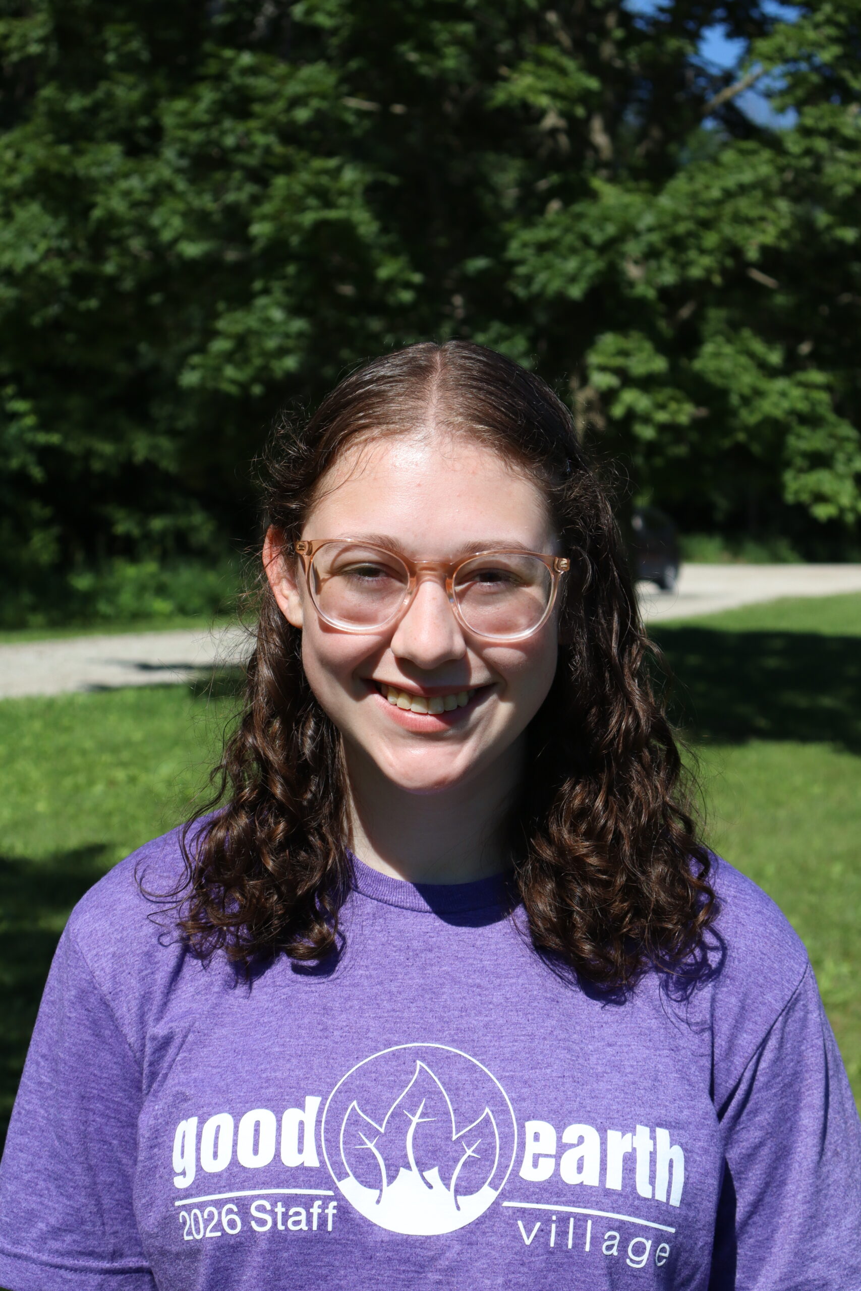 Smiling young woman with wavy brown hair and glasses outdoors in a park; wearing a purple T-shirt that reads 'good earth village 2026 Staff'.