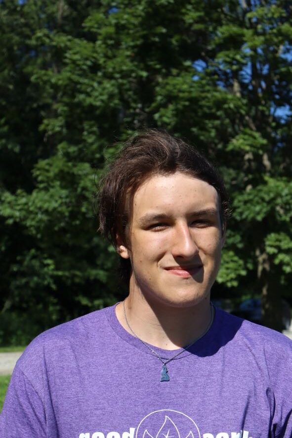 Young man with brown hair wearing a purple shirt, smiling outdoors with green trees in the background.