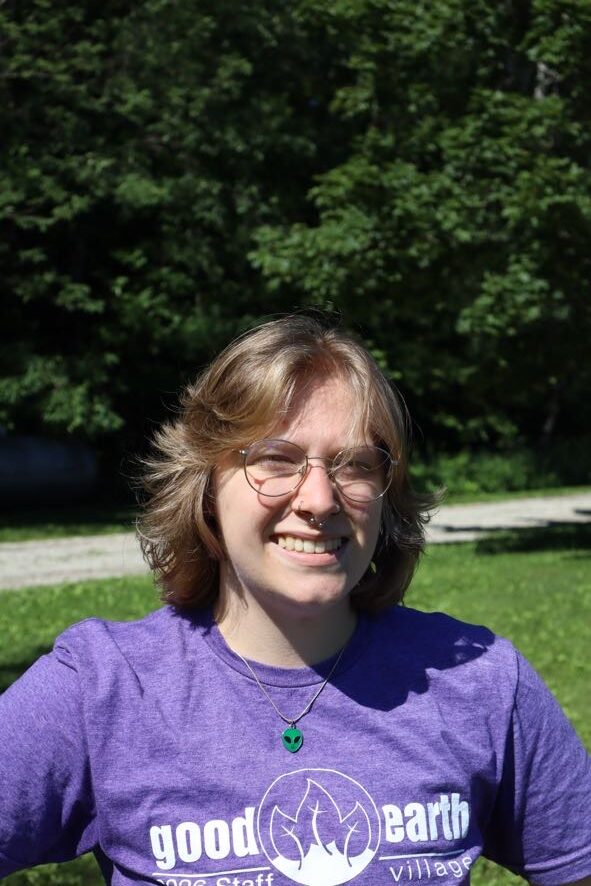 Smiling young woman with short blond hair and glasses outdoors on a sunny day, wearing a purple t-shirt with a 'good earth' logo, trees behind her.