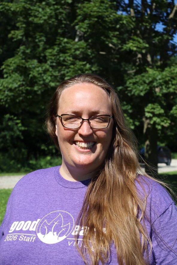 Smiling woman with long hair and glasses outdoors, wearing a purple shirt with a white logo.