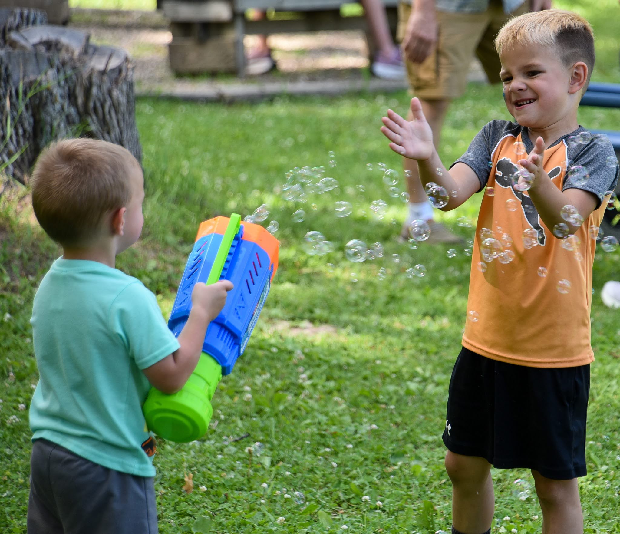 Two young boys outdoors, one aims a blue-and-green bubble blower while bubbles float around them.