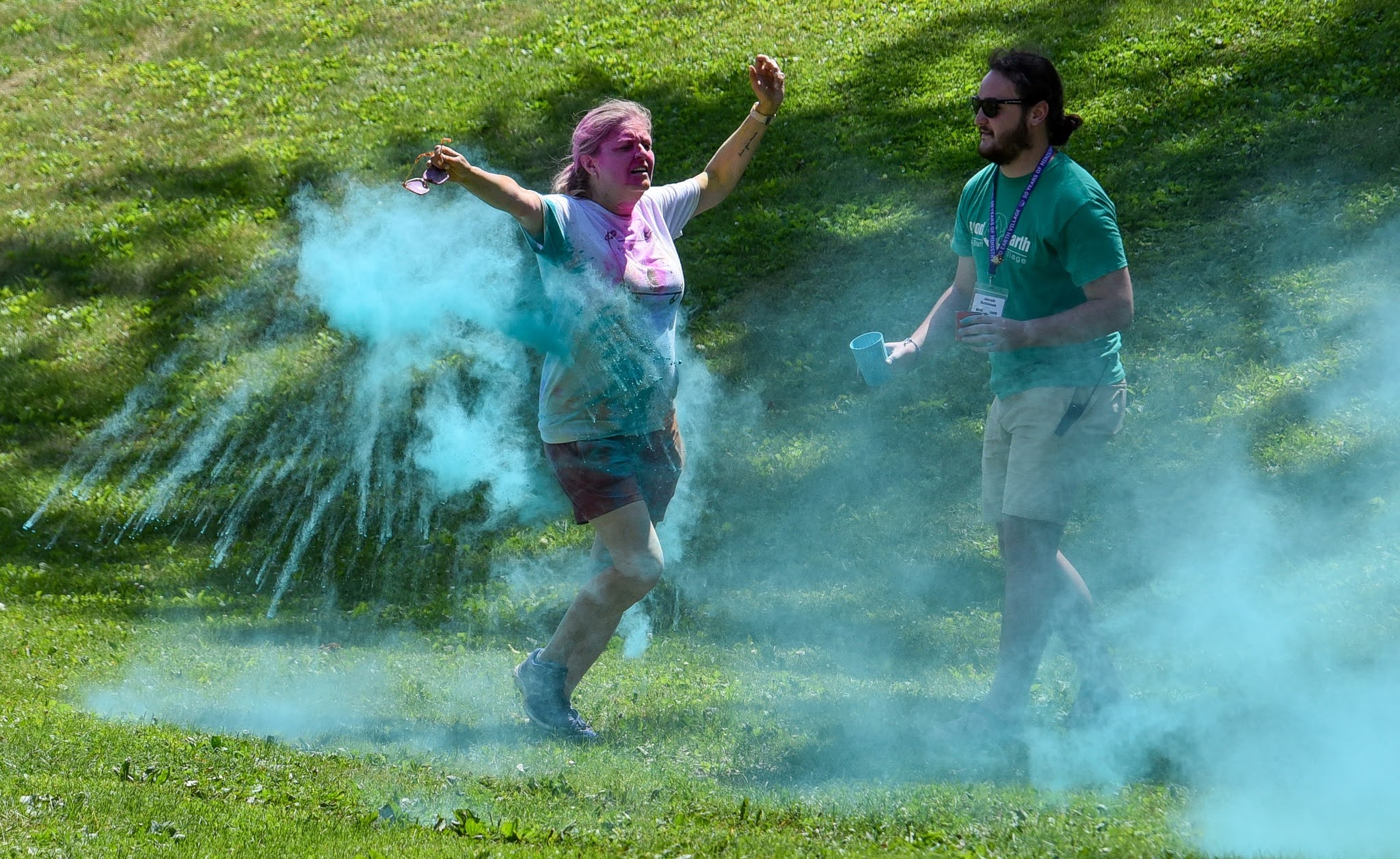 Two people outdoors in a grassy area, one woman flinging blue powder with pink powder on her face while the man watches nearby in sunglasses; blue smoke surrounds them.