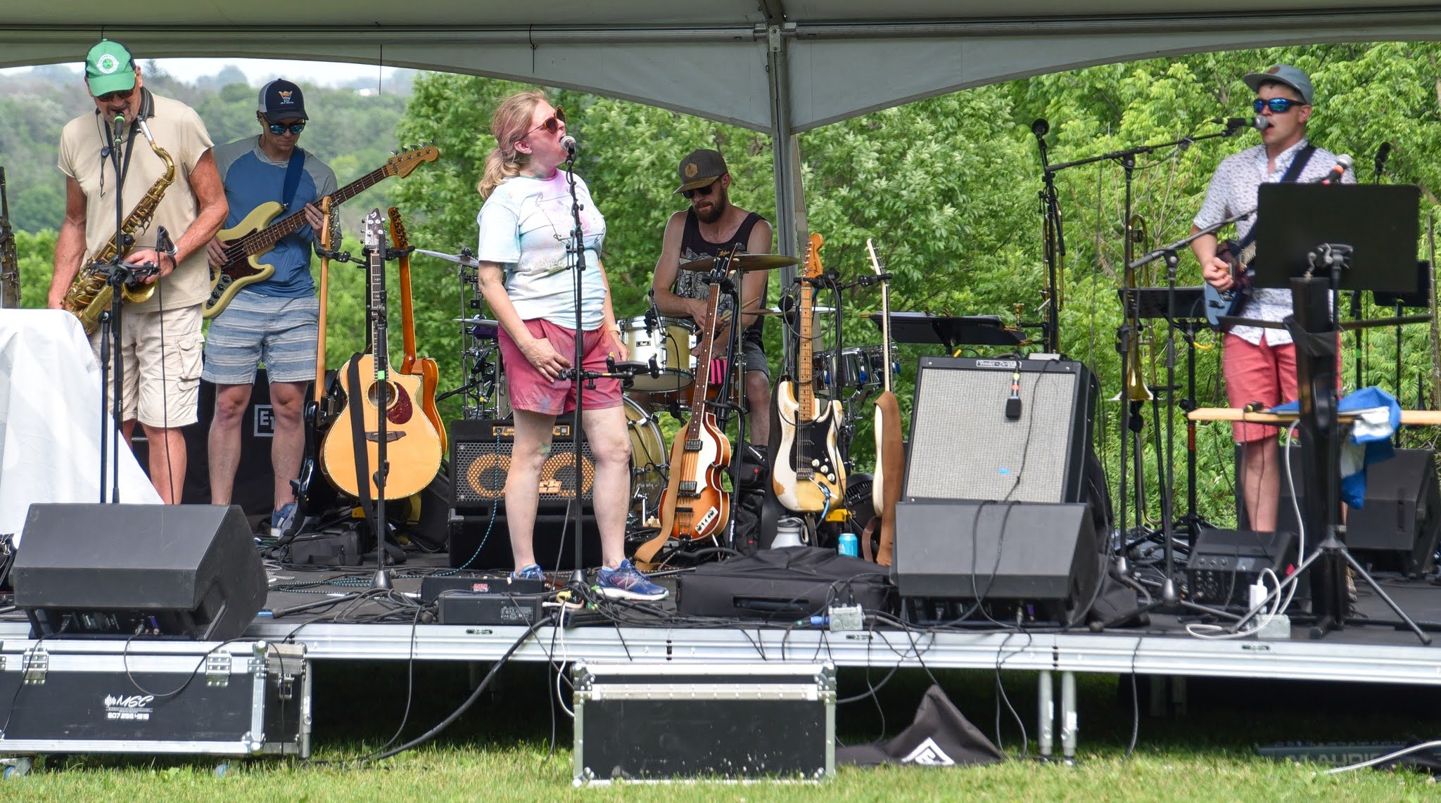 Six-member band performing on an outdoor stage under a canopy, with guitars, drums, and a saxophone player visible behind the lead singer.