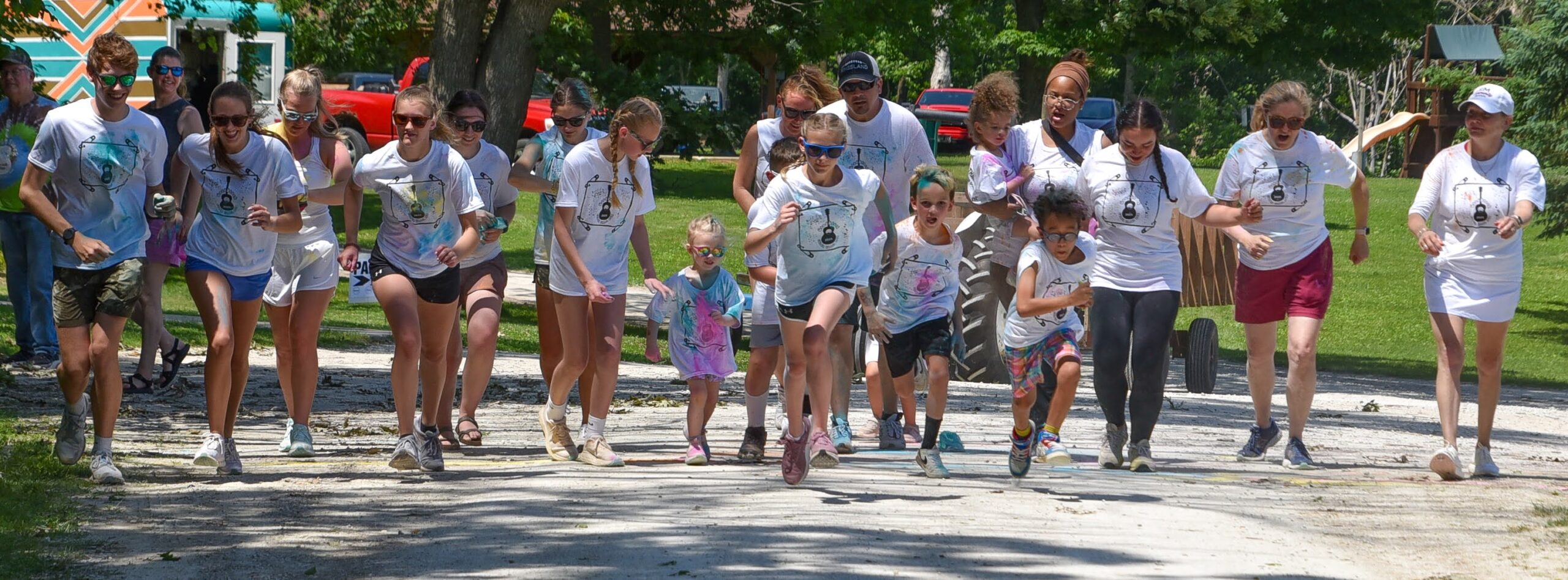 Group of children and adults sprinting together on a sunlit park path in matching white race shirts, starting a run.