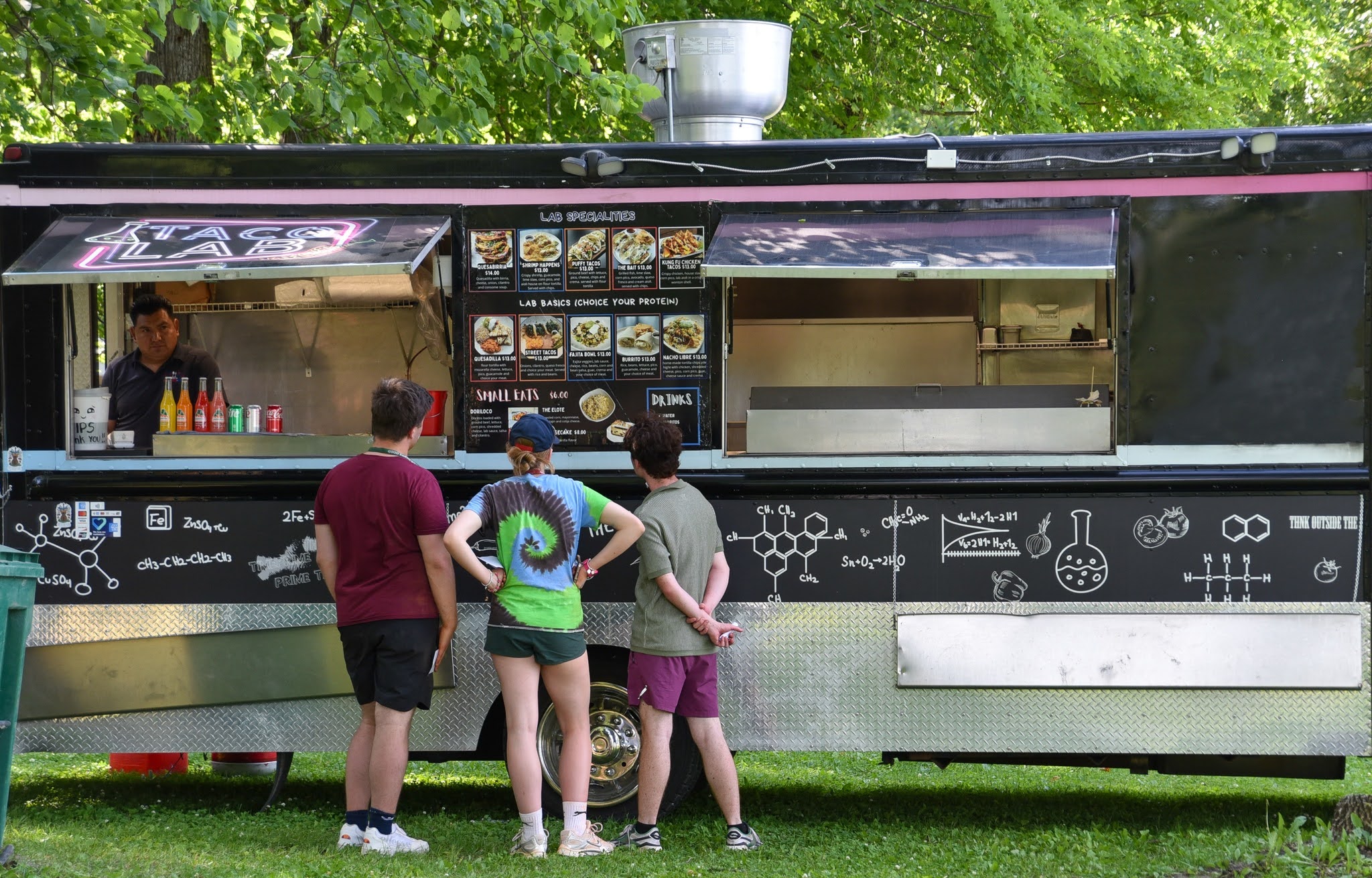 People order food from a black mobile food truck with a lab-themed side panel in a park setting.