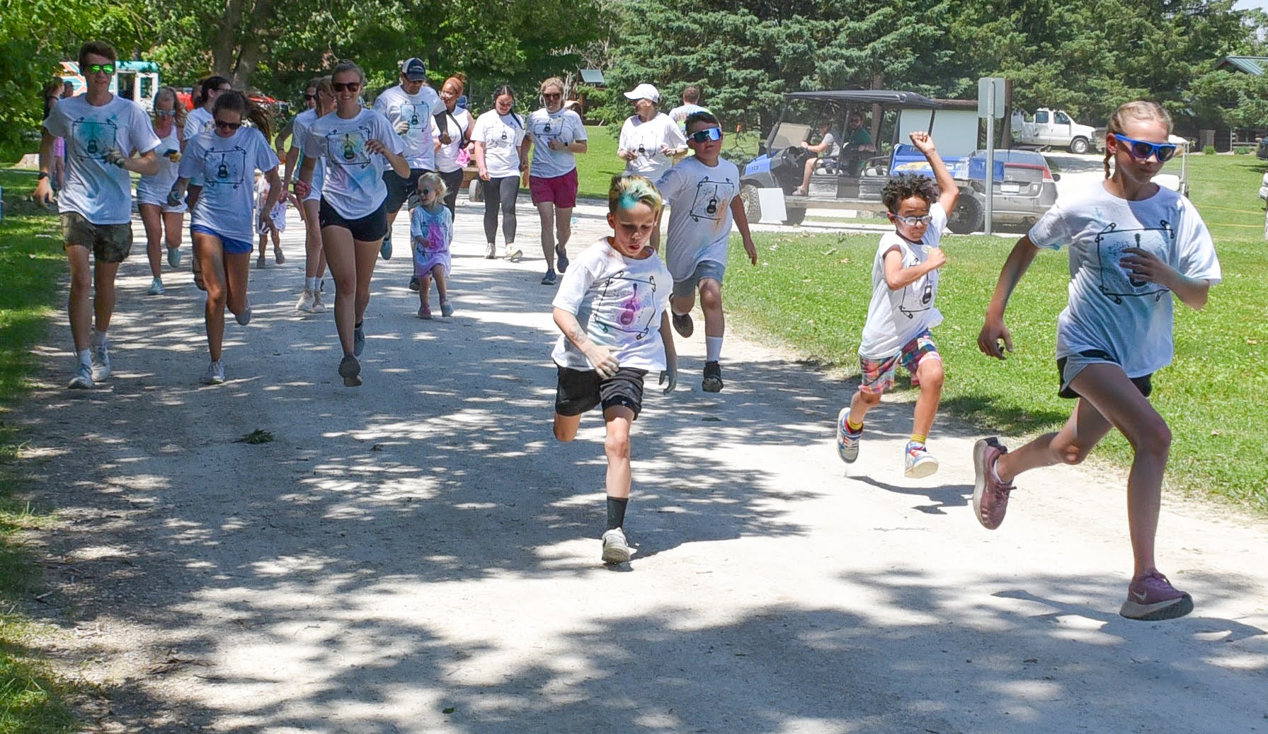 Group of people of various ages running along a sunny park path in matching white t-shirts for a charity race, with trees in the background.