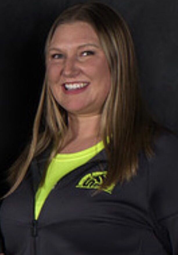Smiling woman with long brown hair, wearing a black zip-front jacket over a neon yellow top, against a dark backdrop.