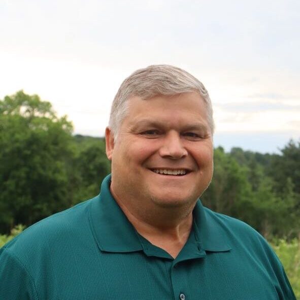 Smiling man with short gray hair wearing a teal polo shirt, outdoors with trees in the background.