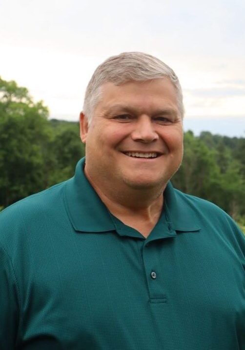 Smiling man with short gray hair wearing a teal polo shirt, outdoors with trees in the background.
