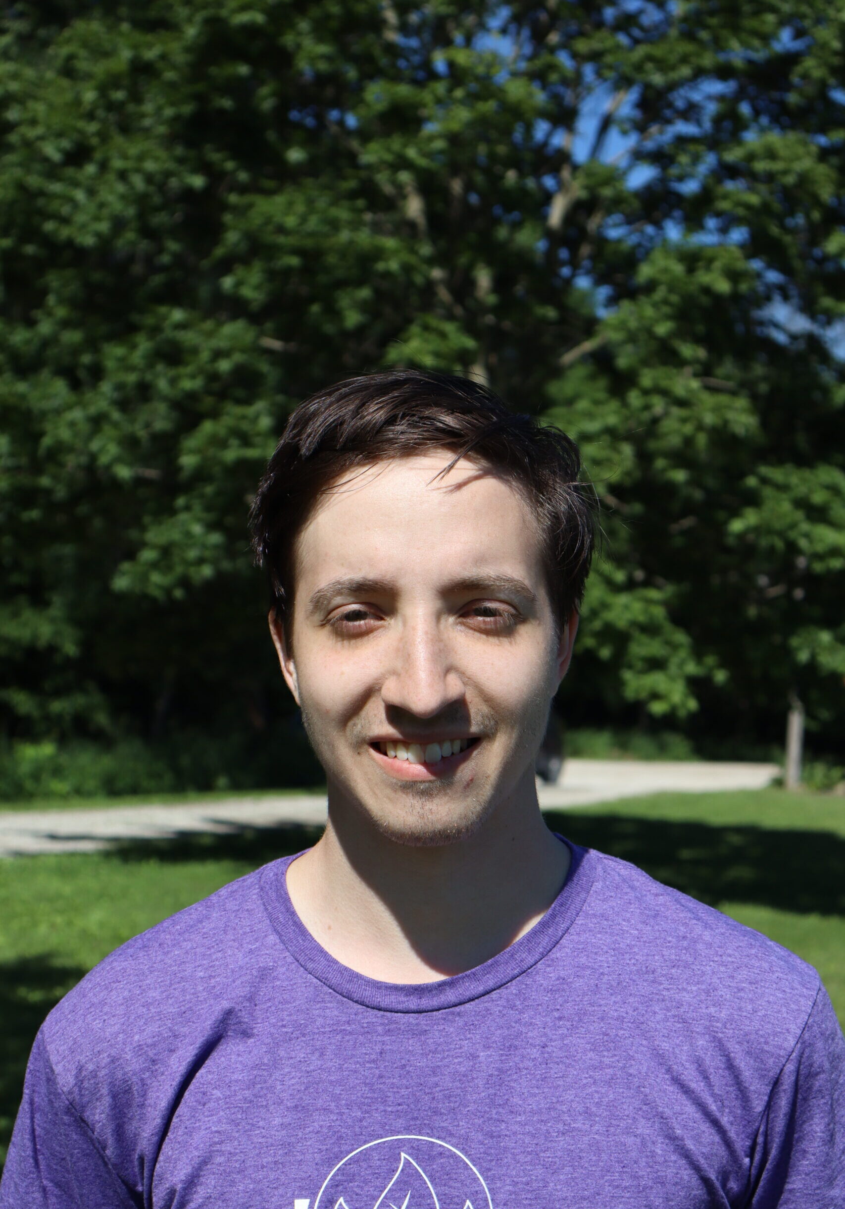 Young man with short dark hair smiling outdoors in a sunny park, wearing a purple shirt.