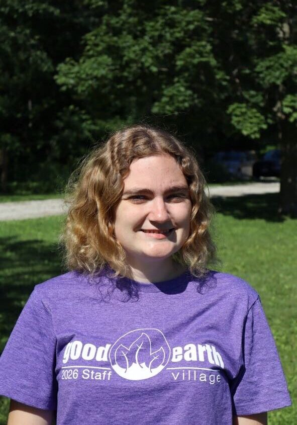 Young woman with shoulder-length wavy blonde hair wearing a purple 'Good Earth Village 2026 Staff' T-shirt, standing on a sunny grassy area with trees in the background.