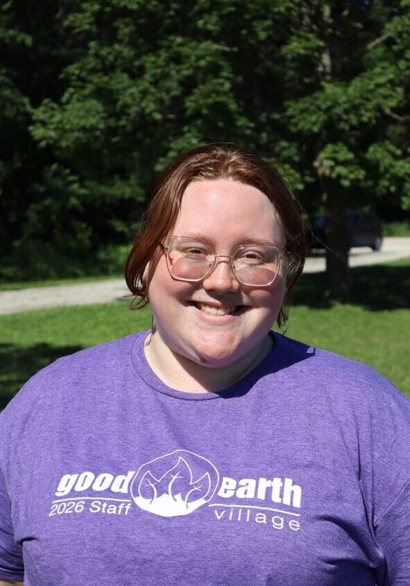 Smiling person with short brown hair and glasses wearing a purple 'Good Earth Village' staff T-shirt outdoors.