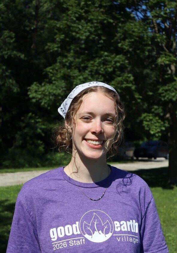 Young woman smiling outdoors in a purple Good Earth Village 2026 Staff shirt and a white headscarf, trees behind.