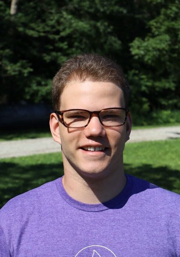 Young man with short brown hair and brown rectangular glasses smiles at the camera, wearing a purple T-shirt outdoors in a sunny park with green trees in the background.