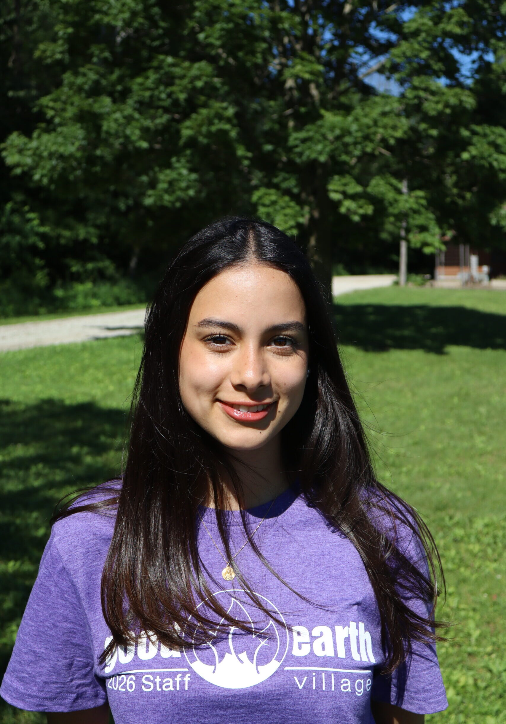 Smiling woman outdoors in a park on a sunny day, wearing a purple shirt with an Earth-themed graphic text.