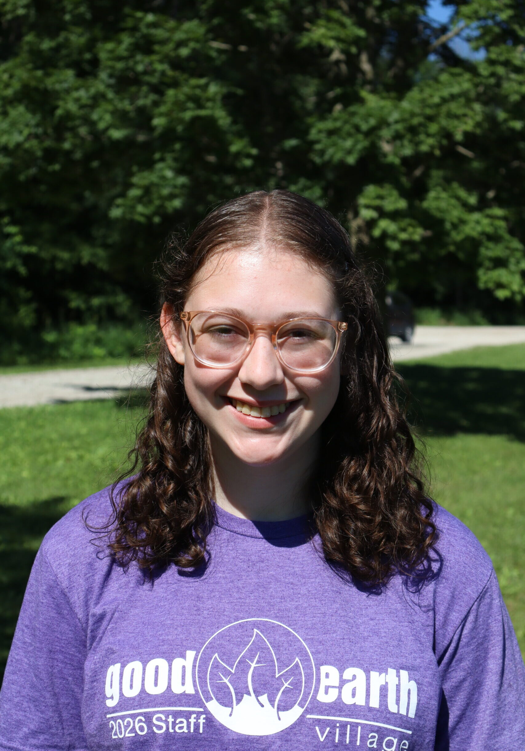 Smiling young woman with wavy brown hair and glasses outdoors in a park; wearing a purple T-shirt that reads 'good earth village 2026 Staff'.