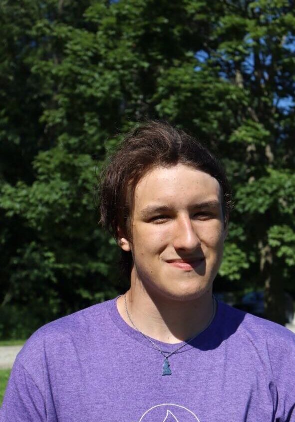 Young man with brown hair wearing a purple shirt, smiling outdoors with green trees in the background.