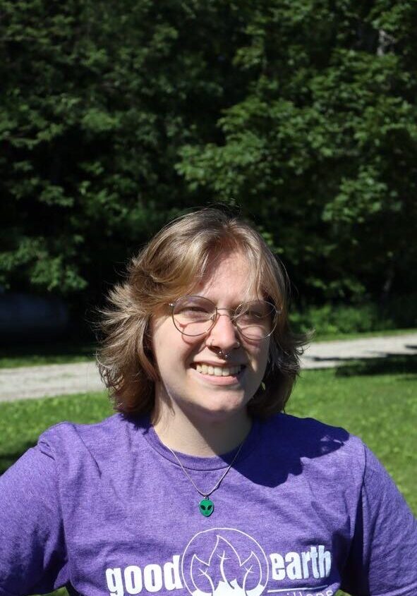 Smiling young woman with short blond hair and glasses outdoors on a sunny day, wearing a purple t-shirt with a 'good earth' logo, trees behind her.