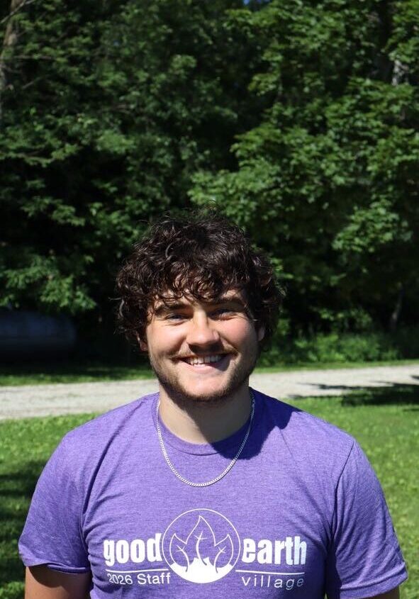 Young man with curly dark hair smiling, wearing a purple Good Earth Village staff shirt, standing outdoors in a sunny grassy area with trees in the background, logo and text visible on his shirt.