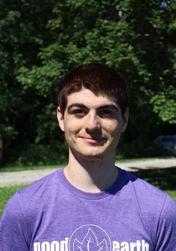 Young man with short dark hair smiling outdoors, wearing a purple 'good earth' T-shirt.