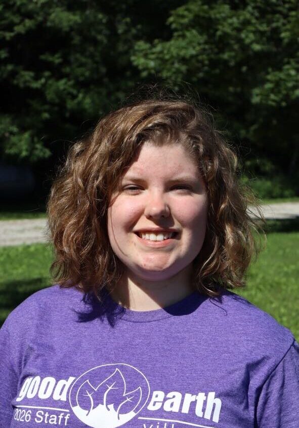 Smiling woman with curly brown hair wearing a purple shirt outdoors on a sunny day, with a 'good earth village' logo visible.