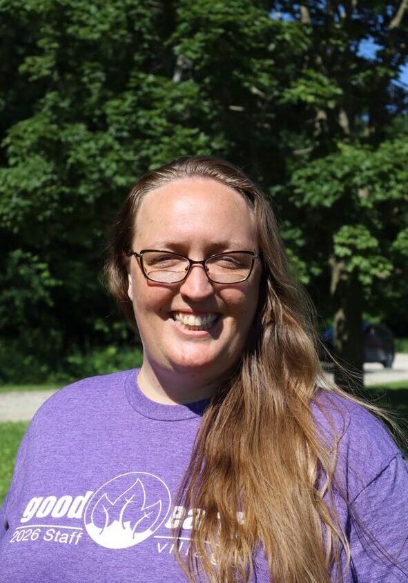 Smiling woman with long hair and glasses outdoors, wearing a purple shirt with a white logo.