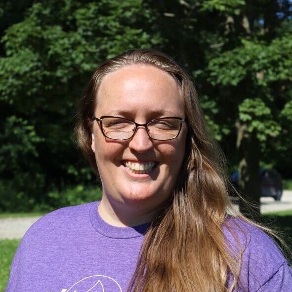 Smiling woman with long hair and glasses outdoors, wearing a purple shirt with a white logo.