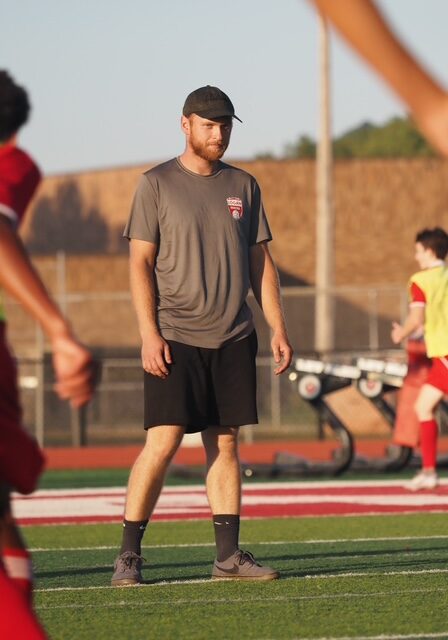 Adult male athlete in a gray t-shirt, black shorts, and cap stands on a sunny athletic field; other players in red jerseys are in the background.
